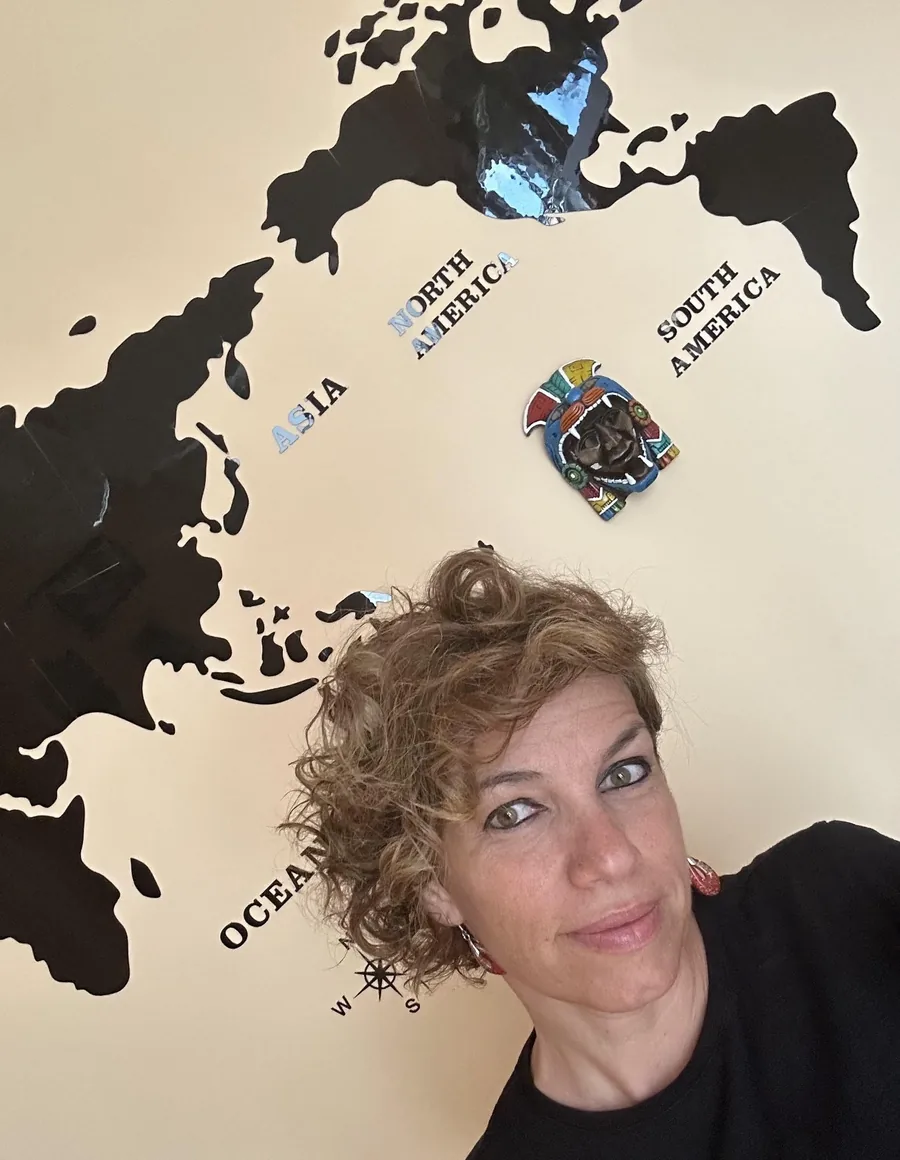 Vincenza Molisano davanti a una grande mappa del mondo nera appesa al muro, con una maschera colorata